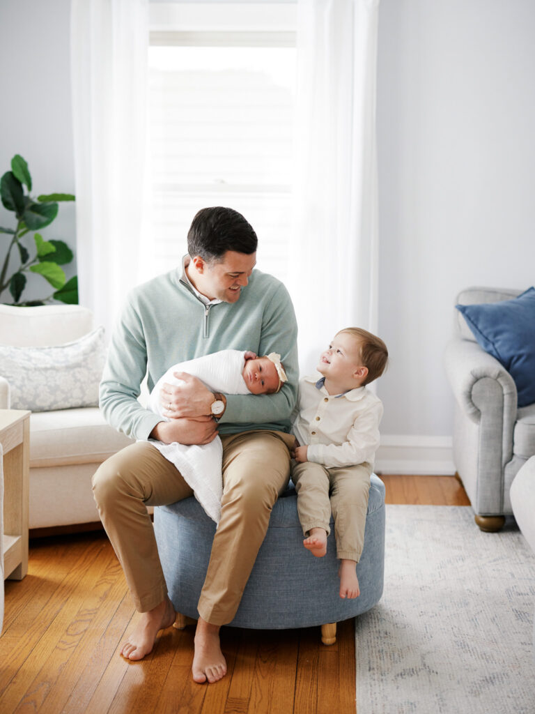 Father seated in blue armchair gazing at newborn baby in lap during in-home session in Western Springs Illinois