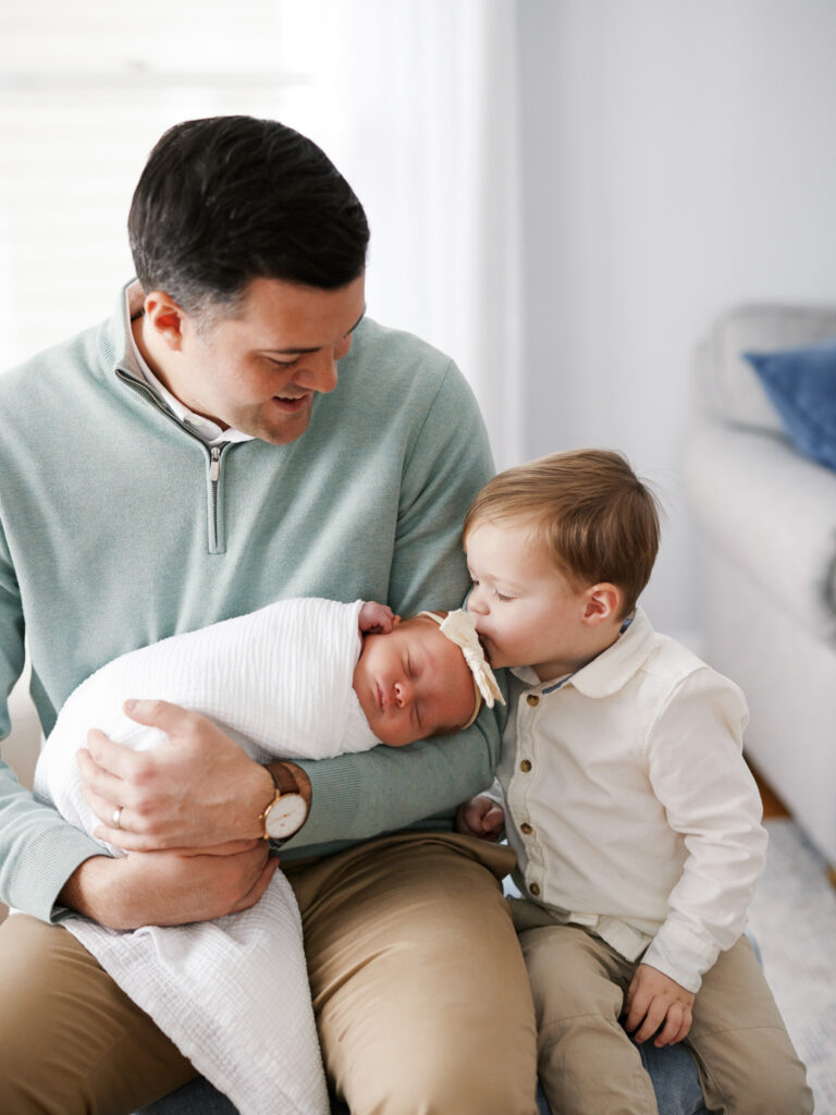 Father seated with toddler sibling both holding newborn baby during in-home newborn session in Western Springs Illinois
