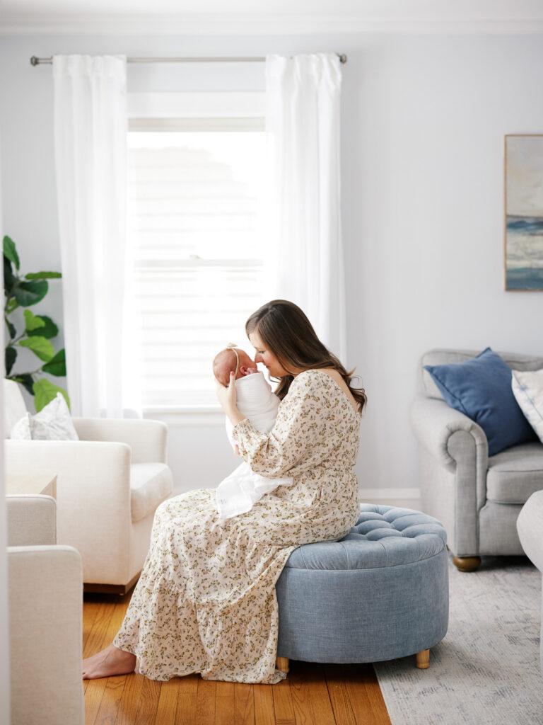 Mother seated on blue ottoman holding sleeping newborn in natural window light during in-home newborn session Western Springs IL