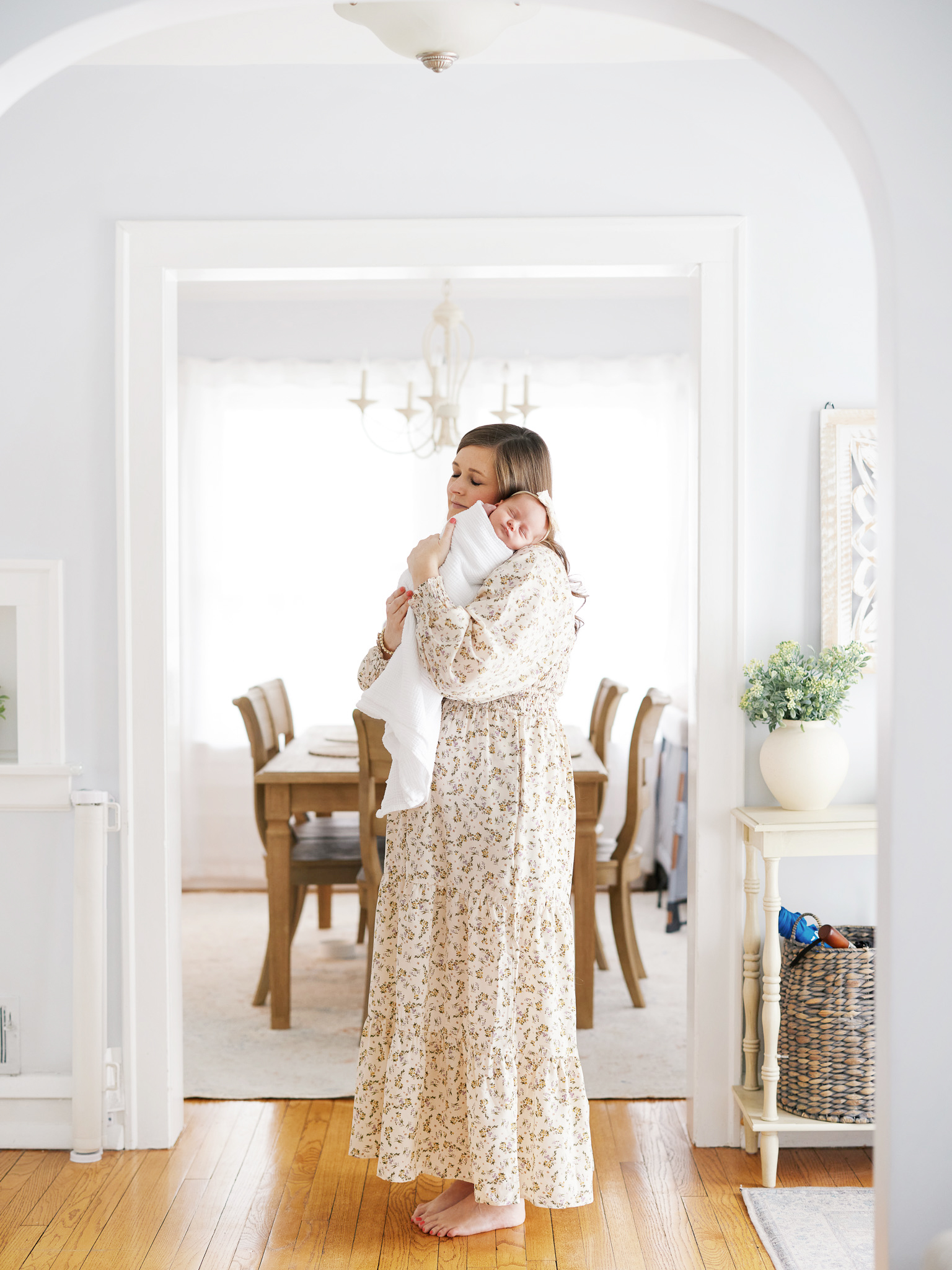Mother in floral dress standing in white arched doorway holding newborn during in-home session in Western Springs Illinois